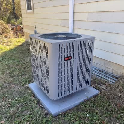 Air conditioning unit installed next to a house, surrounded by grass and trees.